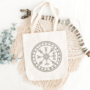 May include: A white canvas tote bag with a brown and gray compass design featuring runes and the text "Vegvisir". The bag is lying on a white surface with a macrame wall hanging behind it.
