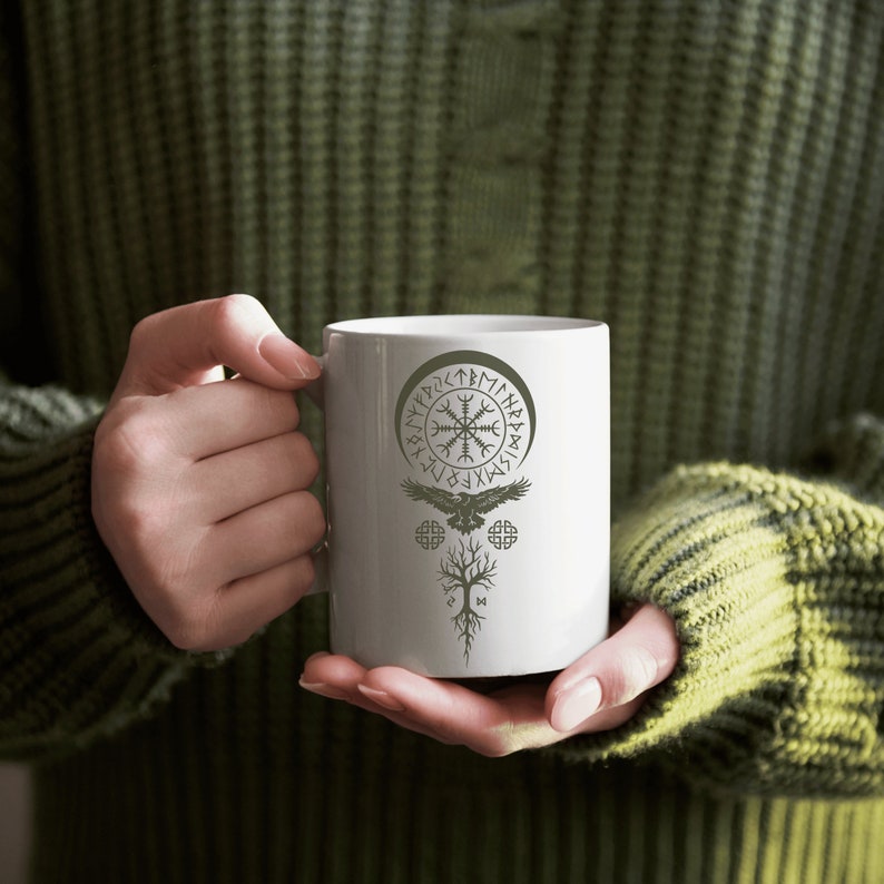 May include: White ceramic mug with a black and white design of a Viking symbol, a tree, and a bird. The symbol is surrounded by a circle of runes.