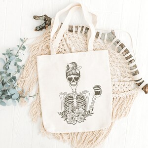May include: A white tote bag with a black and white illustration of a skeleton with a bun in its hair, wearing heart-shaped sunglasses and holding a cup of coffee. The skeleton is surrounded by flowers.