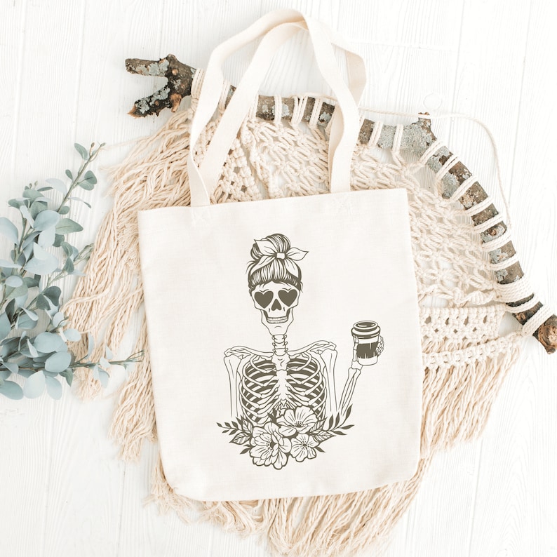 May include: A white tote bag with a black and white illustration of a skeleton with a bun in its hair, wearing heart-shaped sunglasses and holding a cup of coffee. The skeleton is surrounded by flowers.