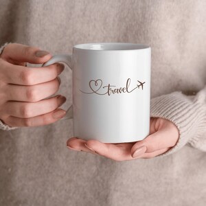 Puede incluir: Taza de cer&aacute;mica blanca con un dise&ntilde;o de coraz&oacute;n marr&oacute;n y avi&oacute;n y la palabra "travel" escrita en cursiva.
