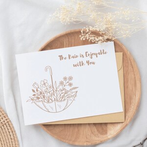 May include: A white card with brown text that reads "The rain is enjoyable with you". A brown line drawing of an umbrella with flowers inside is below the text. The card is on a wooden plate with a white background.