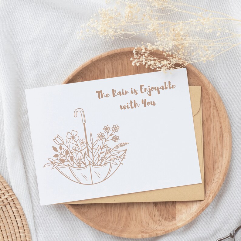 May include: A white card with brown text that reads "The rain is enjoyable with you". A brown line drawing of an umbrella with flowers inside is below the text. The card is on a wooden plate with a white background.