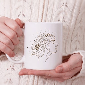 May include: White ceramic mug with a line drawing of a woman's face with planets and stars in her hair.