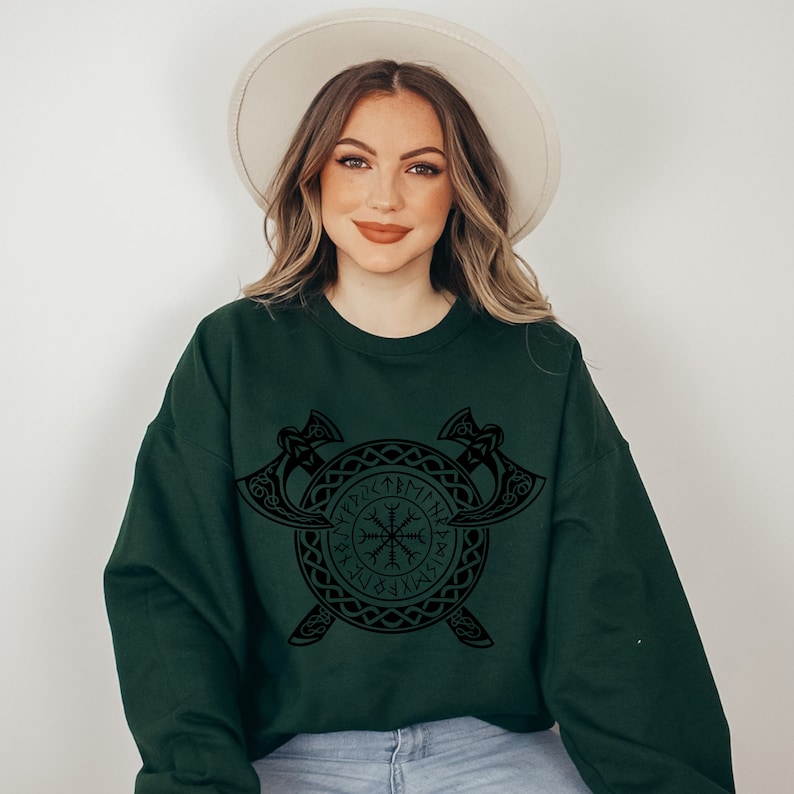K&ouml;nnte beinhalten: Ein dunkelgr&uuml;ner Pullover mit einem schwarzen Design eines Wikinger-Symbols, das von einem Kreis aus Runen und zwei gekreuzten &Auml;xten umgeben ist.