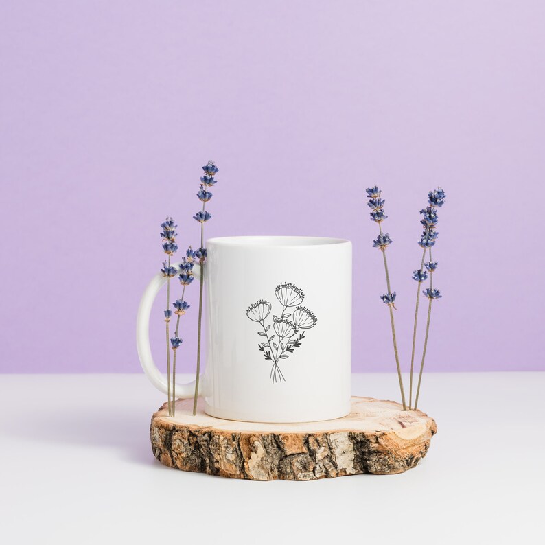 May include: White ceramic mug with a black line drawing of a bouquet of flowers. The mug is sitting on a wooden slice and surrounded by lavender sprigs.