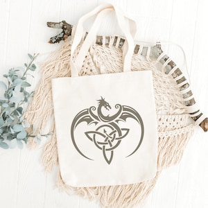 May include: A white canvas tote bag with a gray Celtic knot design featuring a dragon. The design is centered on the bag and is surrounded by a white background.