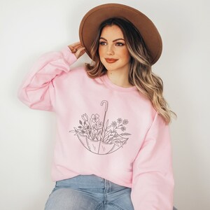 May include: A pink sweatshirt with a graphic of a floral arrangement inside an umbrella. The sweatshirt has a crew neck and long sleeves.