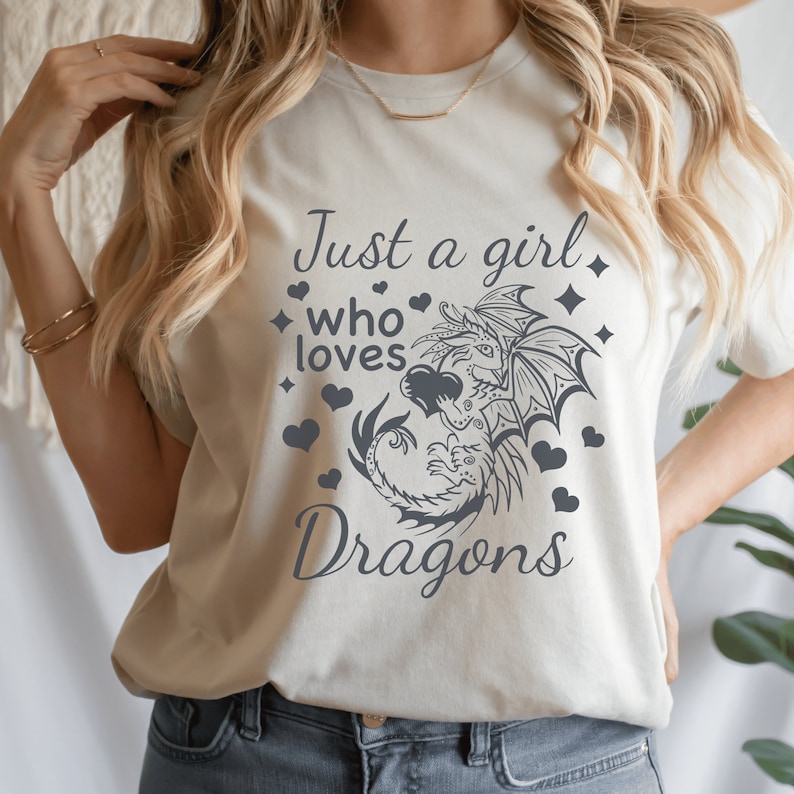 Op de afbeelding: Een wit T-shirt met de tekst "Just a girl who loves Dragons" erop gedrukt. De tekst is in grijs en is omringd door kleine hartjes en sterren. Een gestileerde draak is te zien in het midden van het ontwerp.