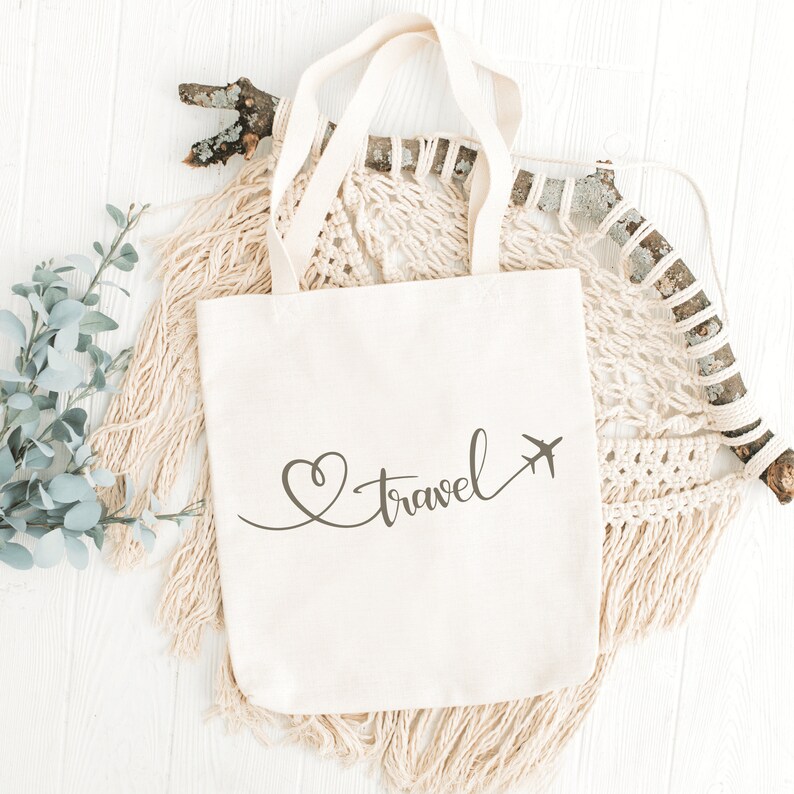 Puede incluir: Una bolsa de lona blanca con el texto "Love Travel" en escritura cursiva con un coraz&oacute;n y un avi&oacute;n gr&aacute;fico.