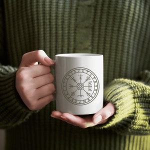 May include: White ceramic mug with a gray compass design and runes around the edge. The mug is being held by a person wearing a green sweater.