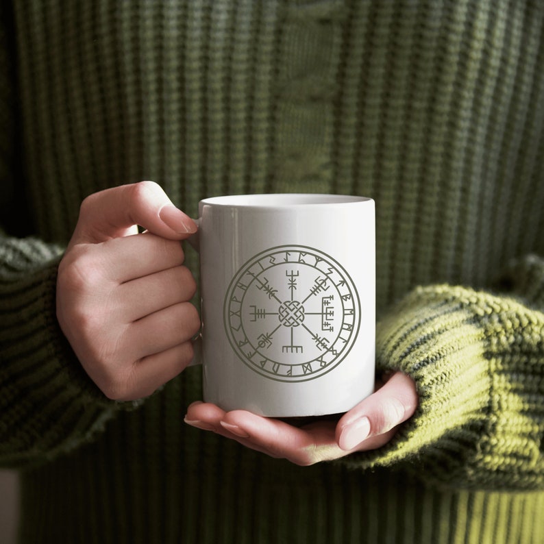 May include: White ceramic mug with a gray compass design and runes around the edge. The mug is being held by a person wearing a green sweater.