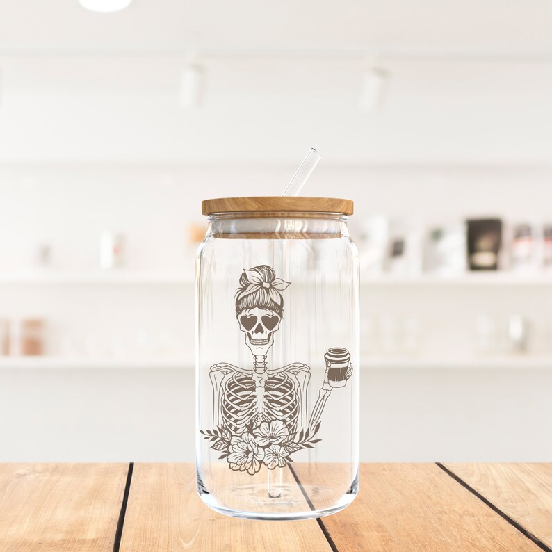 May include: A clear glass tumbler with a wooden lid and a straw. The tumbler has a design of a skeleton with a bun in its hair, wearing sunglasses and holding a cup of coffee. The design is in brown.