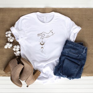 May include: White t-shirt with a brown line drawing of a hand holding a celestial design with a crescent moon, stars, and a planet.