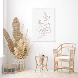 May include: A white framed print of a line drawing of a flower in a brown color, hanging on a white wall. A wicker chair with a white cushion sits in front of the print. A wicker side table with two white ceramic decorative objects sits in front of the chair. A large wicker basket with dried plants sits on the floor.