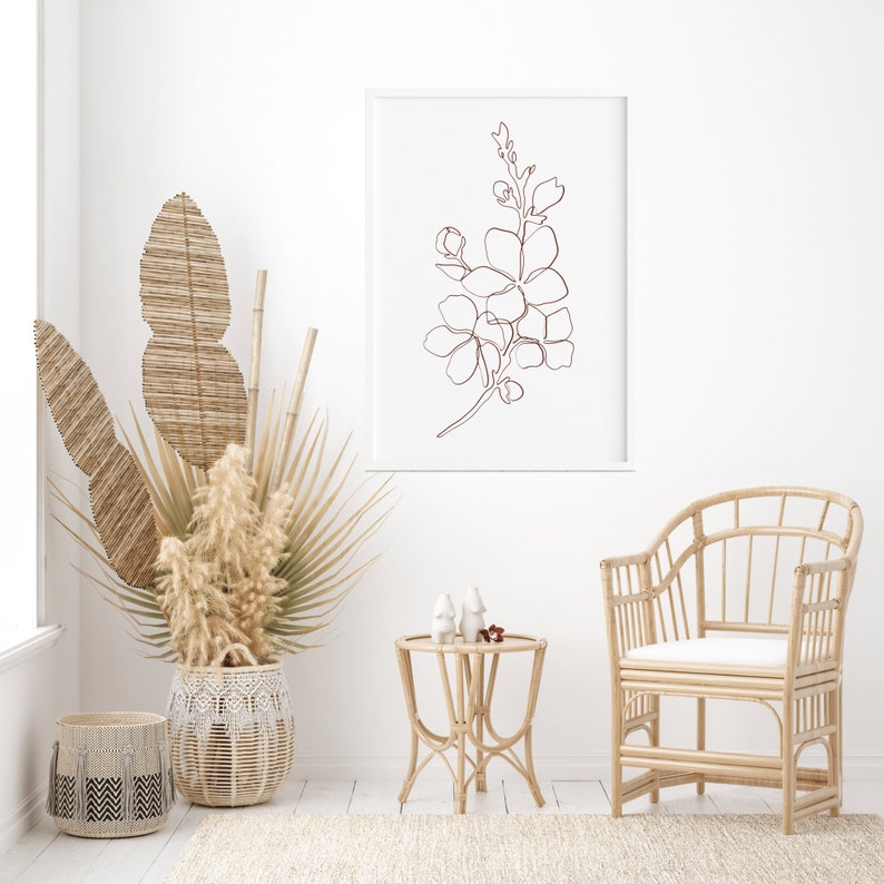 May include: A white framed print of a line drawing of a flower in a brown color, hanging on a white wall. A wicker chair with a white cushion sits in front of the print. A wicker side table with two white ceramic decorative objects sits in front of the chair. A large wicker basket with dried plants sits on the floor.