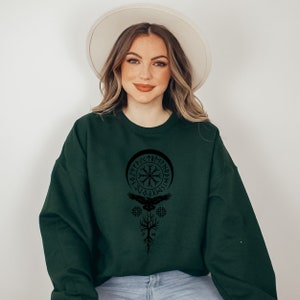 May include: A dark green sweatshirt with a black graphic print of a Viking symbol, a bird, and a tree. The symbol is surrounded by runes.