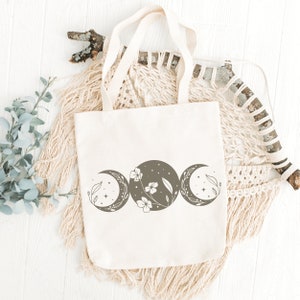 Op de afbeelding: Een witte canvas tote bag met een bruin driedubbel maanontwerp met een bloemenmotief. De tas heeft bruine hengsels en is gefotografeerd op een wit houten oppervlak met een macramee wandhanger.