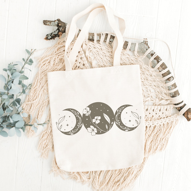 Op de afbeelding: Een witte canvas tote bag met een bruin driedubbel maanontwerp met een bloemenmotief. De tas heeft bruine hengsels en is gefotografeerd op een wit houten oppervlak met een macramee wandhanger.