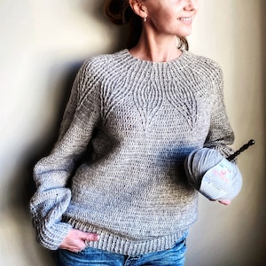 May include: A gray crocheted sweater with a textured pattern. The sweater has a round neckline and long sleeves. The sweater is being held by a person, and a ball of gray yarn is visible in the image.
