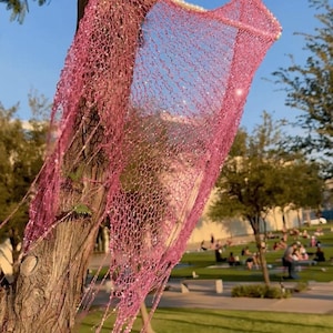 May include: A pink, net-like garment hangs from a tree branch, its delicate structure visible against a blue sky. The garment is adorned with small, sparkling embellishments and hangs from a pearl-studded hanger.