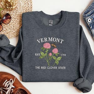 Könnte beinhalten: Ein dunkelgraues Sweatshirt mit dem Wort "VERMONT" in weißem Bogen. Darunter ein rosa Kleeblatt-Design mit einer Biene und dem Text "EST. 1791" und "THE RED CLOVER STATE". Das Sweatshirt ist mit braunen Schuhen und Sonnenbrillen gefaltet.