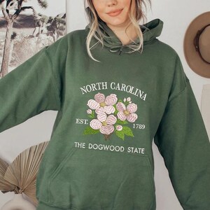 May include: A dark green hoodie with a front pocket and a drawstring hood. The front features an embroidered design with the words "NORTH CAROLINA" above a floral illustration and the text "EST. 1789 THE DOGWOOD STATE".