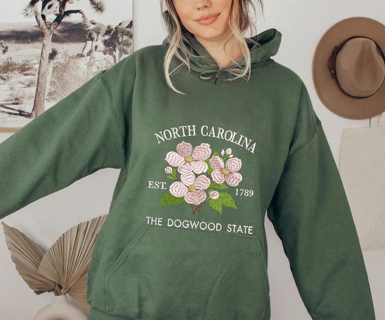 May include: A dark green hoodie with a front pocket and a drawstring hood. The front features an embroidered design with the words "NORTH CAROLINA" above a floral illustration and the text "EST. 1789 THE DOGWOOD STATE".