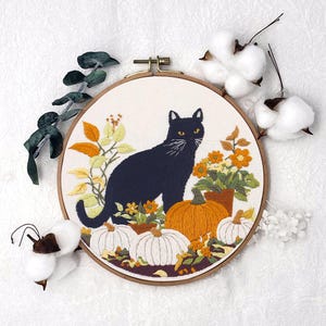 Könnte beinhalten: Gesticktes Bild in einem Holzrahmen, das eine schwarze Katze mit gelben Augen, Kürbissen und Herbstlaub zeigt. Die Katze steht im Mittelpunkt, vor einem cremefarbenen Hintergrund. Kürbisse sind weiß und orange. Der Rahmen hat etwa 20 cm Durchmesser.