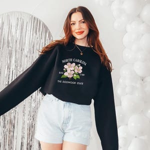 May include: Black sweatshirt with the text "NORTH CAROLINA EST. 1789 THE DOGWOOD STATE" and a floral design. The sweatshirt is paired with light blue denim shorts. White balloons and a silver backdrop are in the background.