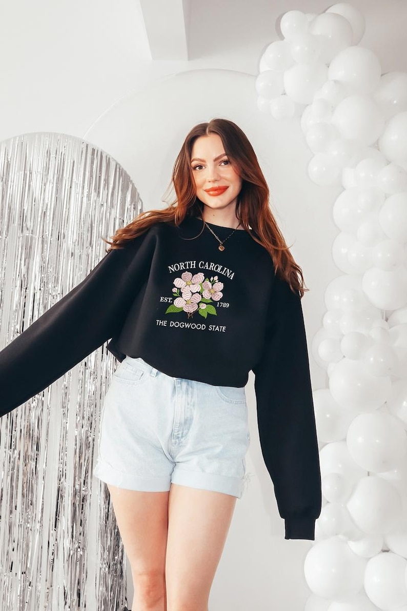 May include: Black sweatshirt with the text "NORTH CAROLINA EST. 1789 THE DOGWOOD STATE" and a floral design. The sweatshirt is paired with light blue denim shorts. White balloons and a silver backdrop are in the background.