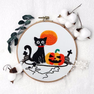 Könnte beinhalten: Stickerei in einem runden Rahmen mit einer schwarzen Katze mit rotem Halsband und einem Kürbis mit grünem Stiel. Der Hintergrund ist weiß mit einer gestickten orangefarbenen Sonne.