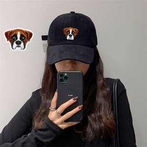 Könnte beinhalten: Eine schwarze Baseballkappe mit einem gestickten braunen und weißen Boxer auf der Vorderseite. Die Kappe hat einen gebogenen Schirm und einen schwarzen verstellbaren Riemen.