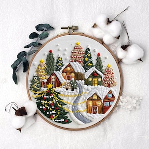 Könnte beinhalten: Gestickte Winterdorf-Szene in einem Holzreifen. Das Kunstwerk zeigt schneebedeckte Häuser, Weihnachtsbäume und einen gewundenen Pfad. Die Farbpalette umfasst Weiß, Grün, Rot und Gelb und schafft ein festliches Urlaubs-Thema.