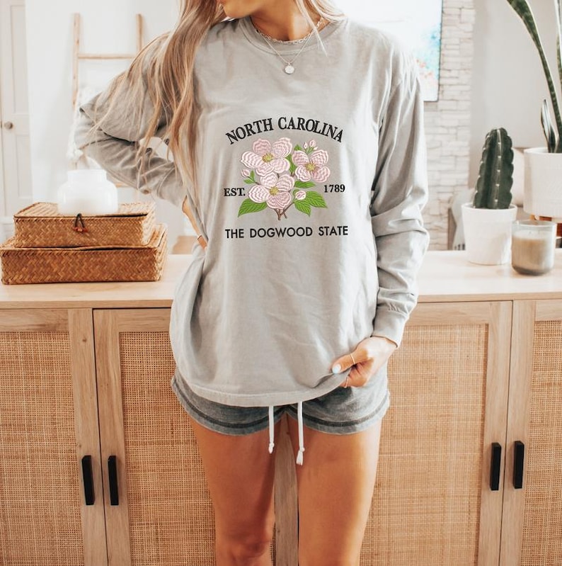 May include: A long-sleeved, light gray shirt featuring the text "North Carolina" above a floral design and the words "Est. 1789" and "The Dogwood State". The shirt is paired with gray shorts.