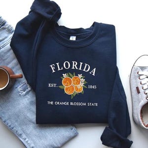 Könnte beinhalten: Ein marineblaues Sweatshirt mit dem Wort "FLORIDA" in weißer Schrift, einem Orangenblüten-Design und dem Text "EST. 1845" und "THE ORANGE BLOSSOM STATE". Das Sweatshirt wird mit Jeans und grauen Turnschuhen kombiniert.