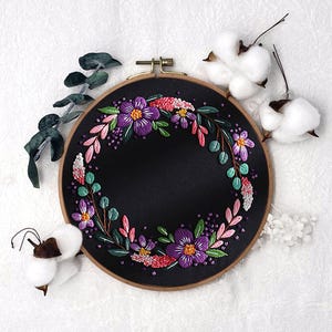 Könnte beinhalten: Ein schwarzer Stickrahmen mit einem floralen Kranzdesign. Der Kranz besteht aus gestickten lila, rosa und grünen Blumen und Blättern. Der Rahmen ist aus Holz und hat einen messingfarbenen Verschluss. Baumwollkapseln und Eukalyptuszweige umgeben den Rahmen.