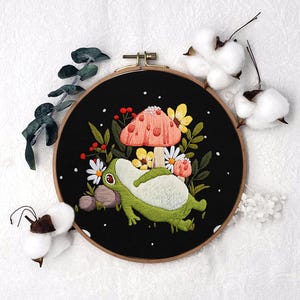 Könnte beinhalten: Gesticktes Stickrahmen-Kunstwerk mit einer grünen und weißen Frosch-Illustration neben einem pfirsichfarbenen Pilz mit roten Punkten und Blumen. Der Hintergrund ist schwarz, und der Holzrahmen hat einen Durchmesser von ca. 20 cm.