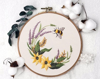 Peut inclure: Broderie sur tambour représentant une abeille et des fleurs. Le motif comprend des tournesols jaunes, de la lavande violette et des feuilles vertes sur un tissu crème. Le tambour en bois est visible. Thème : nature et décoration intérieure.