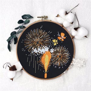 Könnte beinhalten: Ein schwarzer Stickrahmen mit einem Motiv aus Feuerwerk in orange, gelben und weißen Fäden. Der Rahmen ist von weißen Wattebäuschen und grünen Eukalyptusblättern umgeben.