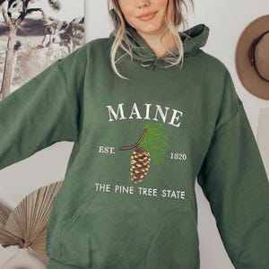Könnte beinhalten: Dunkelgrüner Kapuzenpullover mit dem Wort "MAINE" in Weiß, einer Kiefernzapfen-Grafik und dem Text "THE PINE TREE STATE". Der Hoodie hat eine Kapuze mit Kordelzug und eine Vordertasche. Der Text "EST. 1820" ist ebenfalls enthalten.