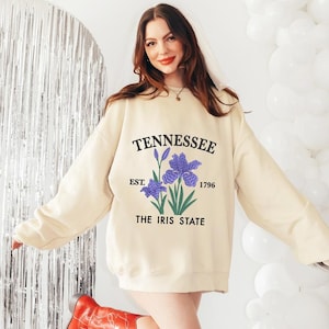 Puede incluir: Sudadera color crema con la palabra "TENNESSEE" sobre un gráfico de flor de iris púrpura. Debajo, las palabras "EST. 1796" y "THE IRIS STATE". La persona lleva botas de vaquero rojas.