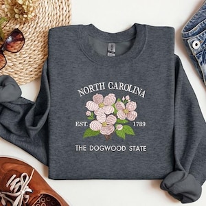 May include: Dark gray sweatshirt with the text "NORTH CAROLINA" and "THE DOGWOOD STATE". The sweatshirt features an embroidered floral design with pink flowers and green leaves. The year "1789" is also included in the design.