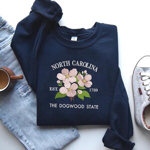 May include: Navy blue sweatshirt with embroidered dogwood flowers and text reading "North Carolina Est. 1789 The Dogwood State." The sweatshirt is paired with light wash jeans, a coffee mug, and gray sneakers.