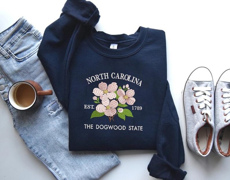 May include: Navy blue sweatshirt with embroidered dogwood flowers and text reading "North Carolina Est. 1789 The Dogwood State." The sweatshirt is paired with light wash jeans, a coffee mug, and gray sneakers.