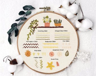 May include: A close-up of a round embroidery hoop with various embroidery stitches and their names. The stitches are worked in different colors of thread, including green, yellow, orange, and red. The hoop is on a white background.
