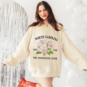 May include: Cream-colored sweatshirt with the text "NORTH CAROLINA" above a floral design and "EST. 1789 THE DOGWOOD STATE" below. The sweatshirt is worn with red cowboy boots.