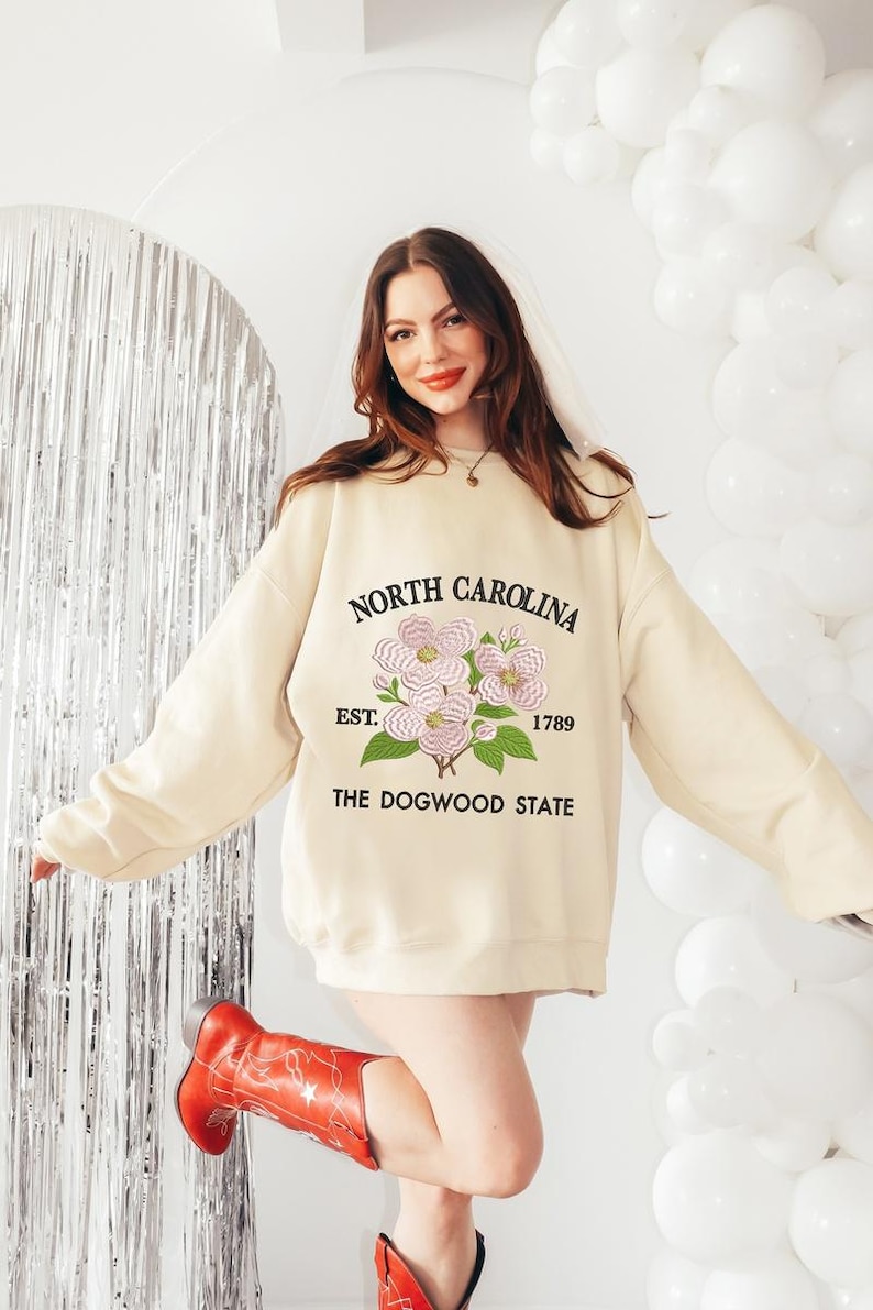 May include: Cream-colored sweatshirt with the text "NORTH CAROLINA" above a floral design and "EST. 1789 THE DOGWOOD STATE" below. The sweatshirt is worn with red cowboy boots.