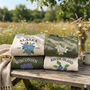 May include: Four sweatshirts stacked on a wooden table, each with state names and floral embroidery. The sweatshirts are in cream and olive green, with the states of Alaska, Michigan, Wisconsin, and Rhode Island displayed.