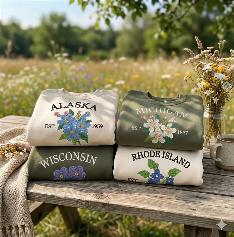May include: Four sweatshirts stacked on a wooden table, each with state names and floral embroidery. The sweatshirts are in cream and olive green, with the states of Alaska, Michigan, Wisconsin, and Rhode Island displayed.